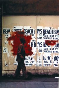 Photo of Richard Nagler's photograph "Beach Boys." Artwork depicts a man walking in front of plywood that has partially shredded Beach Boys concert street posters on it. Wall and posters have been defaced with red paint.