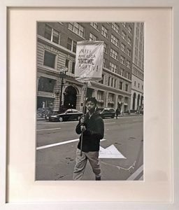 Photo of Richard Nagler's photograph "Make America Think Again!" Artwork depicts a young man walking down the street carrying the sign "Make America Think Again!"