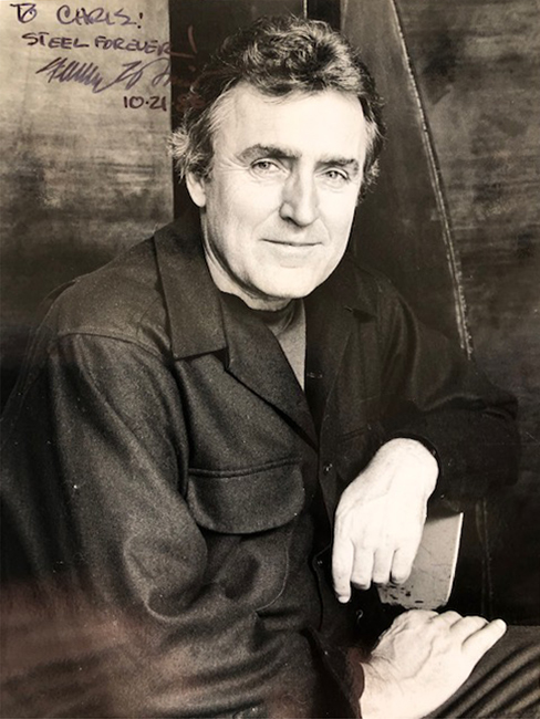 Digital image of Chris Felver's photograph of Fletcher Benton. Image depicts Benton sitting with both arms bent. Photograph has been signed "To Chris, Steel Forever" and signed by Fletcher Benton, dated 10-21-86..