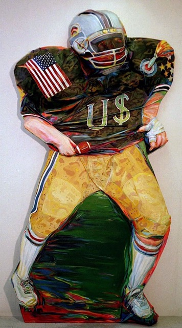 Photo of Diana Krevsky's sculptured painting "Offensive Play." Image depicts a football player in a helmet, brown jersey and gold pants. The jersey has an american flag on one shoulder, a rocket hitting a target on the other, with a large US in the center front (the "S" is made of a $ symbol).