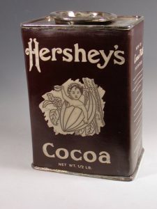 Karen Shapiro ceramic representation of a Hershey's Cocoa Tin. It has a brown background with a white text logo, a figure, and silver cap.
