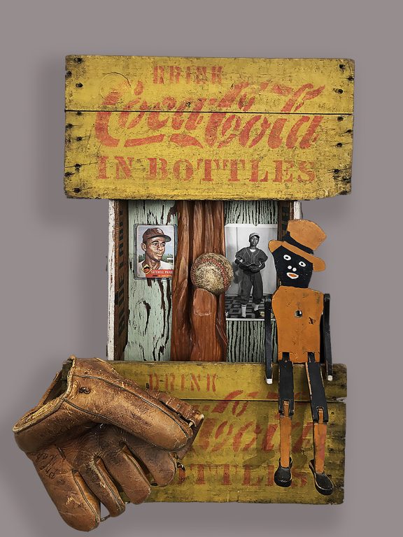 Sculpture of hand carved wood hand - holding an old baseball framed in an old Coke box with vintage Coke signs, old original baseball card of Satchel Paige and original vintage photo of young Black boy in baseball uniform, vintage glove and “dancing Dan” wood toy signifying the Jim Crow era. Vintage advertising tins on the sides of the box.