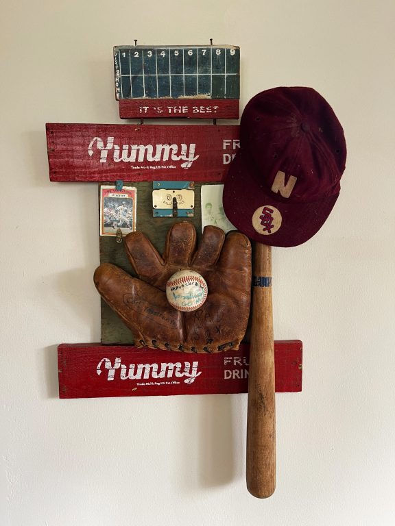 Kat Flyn's sculpture "Carl Yastrzemski I Love You" made from vintage wood backboard framed with Yummy Drink box sides, Old scoreboard on top, vintage baseball glove with boy's handwritten Carl Yastrzemski autograph, Red Sox logo and old Sox baseball hat, old bat, early baseball card of Carl Yastrzemski, vintage score clicker, old faded photo of boys in baseball uniforms.