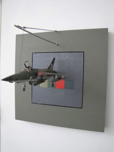 Sculpture of a model jet protruding from a gray, green and red square backdrop.