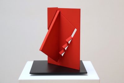 Red metal sculpture of a rectangle with a protruding corner of a triangle and cut out zig zag pattern in the center