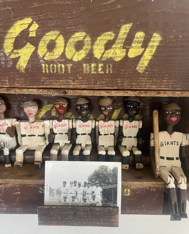 Vintage Goody soft drink box fashioned into a dugout with hand carved & painted wood kid baseball players and an adult BB player inside. Surrounding the dugout are vintage baseball artifacts including a tin baseball game, vintage Willie Mays picture book from a school library, vintage Willie Mays baseball glove and a pullout drawer at the bottom containing an original photo of Black & white kids in baseball uniforms and a vintage well loved Dave Pope baseball card.
